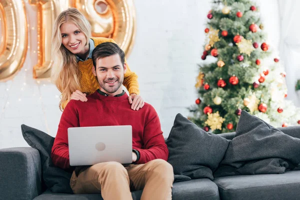 Улыбающаяся пара с помощью ноутбука вместе дома с рождественской елкой — стоковое фото