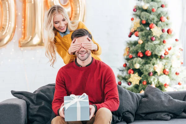 Chica cerrando los ojos y haciendo sorpresa con caja de regalo para novio en casa con árbol de Navidad - foto de stock
