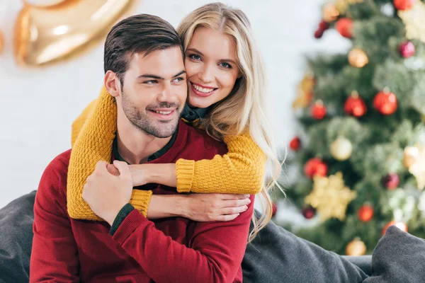 Улыбающаяся девушка обнимает своего парня дома рождественской ёлкой — стоковое фото