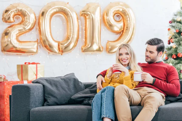 Junges Paar mit Kaffeetassen zu Hause mit goldenen Luftballons für das neue Jahr 2019 — Stockfoto