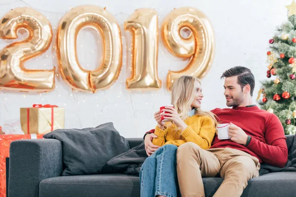 Pareja sosteniendo tazas de café y mirándose en casa con 2019 globos de oro año nuevo - foto de stock