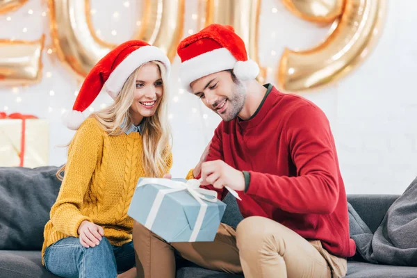 Jeune couple en chapeaux de Père Noël avec cadeau de Noël à la maison avec 2019 ballons dorés — Stock Photo