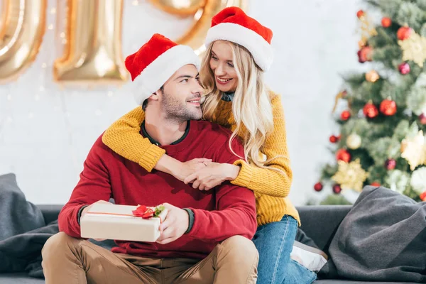 Pareja feliz en sombreros de Santa con regalo de Navidad en casa - foto de stock