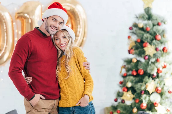 Glückliches junges Paar in Weihnachtsmützen, das sich zu Hause mit Weihnachtsbaum umarmt — Stockfoto