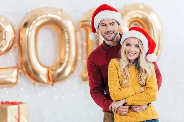 Красивая улыбающаяся пара в шляпах Санта-Клауса обнимается дома с золотыми шариками 2019 года — стоковое фото