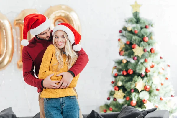 Homem feliz abraçando sua namorada sorridente em casa com árvore de natal — Fotografia de Stock