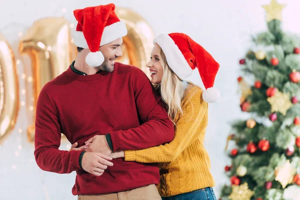 Jovem casal em santa chapéus abraçando e olhando uns para os outros em casa com árvore de natal — Fotografia de Stock