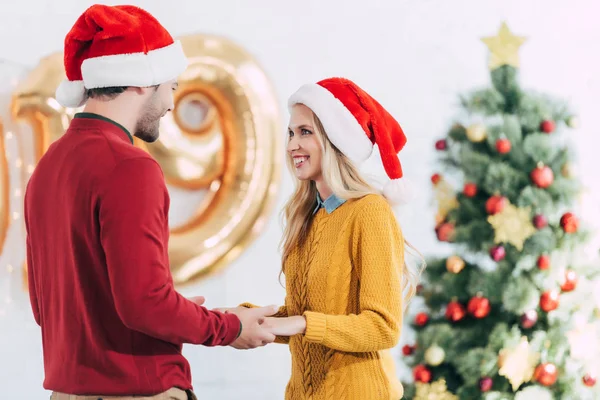 Feliz pareja en sombreros de santa cogidos de la mano y mirándose el uno al otro en la víspera de Navidad - foto de stock