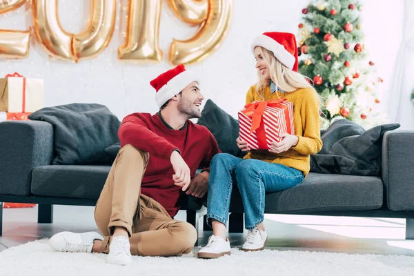 Paar in Weihnachtsmützen mit Weihnachtsgeschenk sitzt auf Sofa — Stockfoto