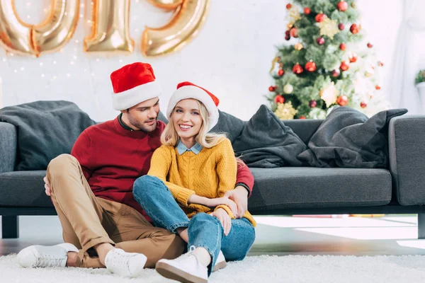 Coppia sorridente in cappelli di Babbo Natale che abbraccia e siede sul pavimento a casa con albero di Natale — Foto stock