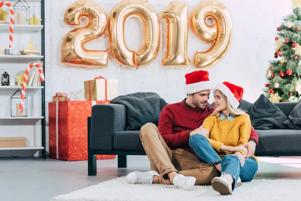Glückliches Paar in Weihnachtsmützen, das sich umarmt und zu Hause mit goldenen Luftballons zum neuen Jahr 2019 auf dem Boden sitzt — Stockfoto