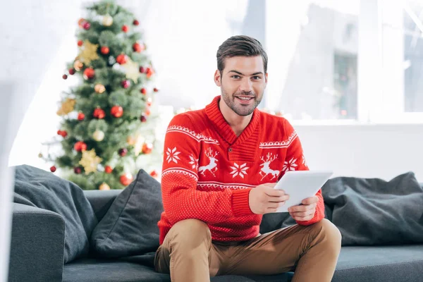 Happy man using digital tablet and sitting on sofa with christmas tree behind — Stock Photo
