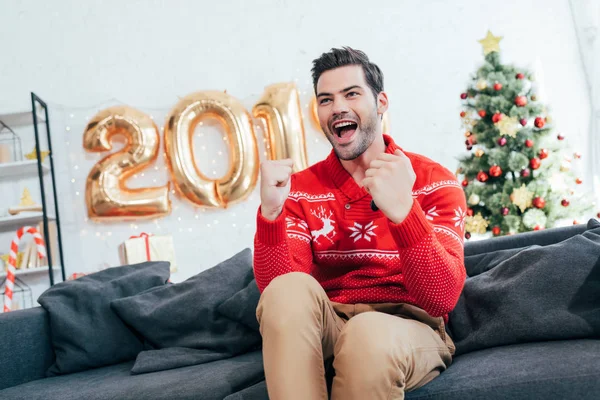 Vencedor animado sentado no sofá com balões e árvore de Natal atrás — Fotografia de Stock