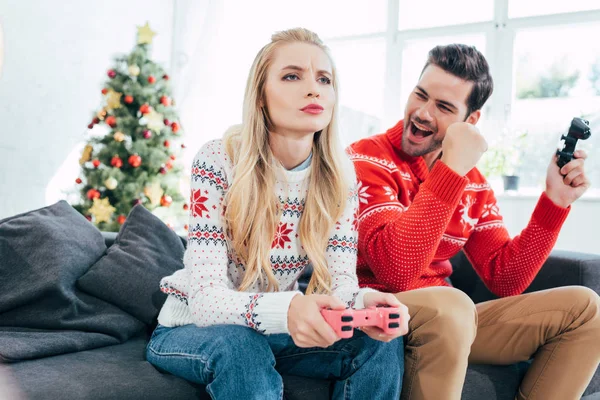 Young couple playing video game with joysticks at home with christmas tree — Stock Photo