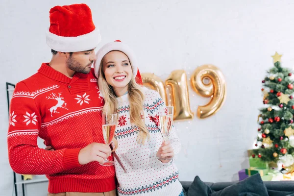 Coppia in cappelli di Babbo Natale che tiene bicchieri di champagne e abbraccia a casa con i palloncini di Capodanno 2019 — Foto stock