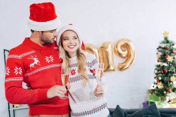 Pareja en sombreros de santa celebración de copas de champán y abrazos en casa con 2019 globos de año nuevo - foto de stock