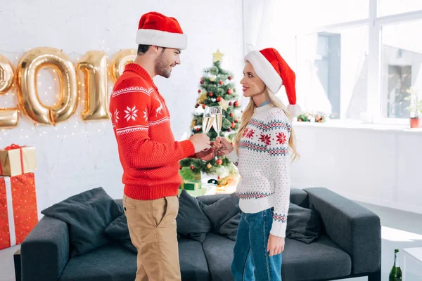 Beautiful couple in santa hats clinking with champagne glasses at home with christmas tree — Stock Photo