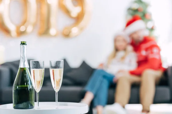 Foco seletivo de garrafa de champanhe e copos na mesa para o ano novo, casal sentado atrás — Fotografia de Stock