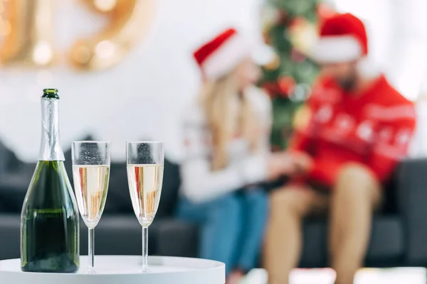 Enfoque selectivo de botella de champán y vasos para celebrar la Navidad, pareja sentada detrás - foto de stock