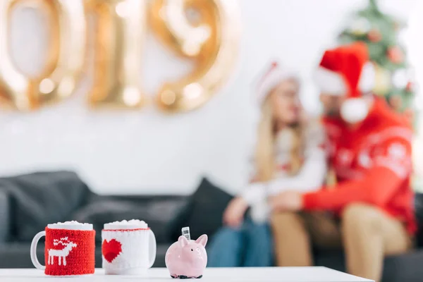 Focus selettivo di tazze di caffè natalizie e salvadanaio con banconote sul tavolo, coppia seduta dietro — Foto stock