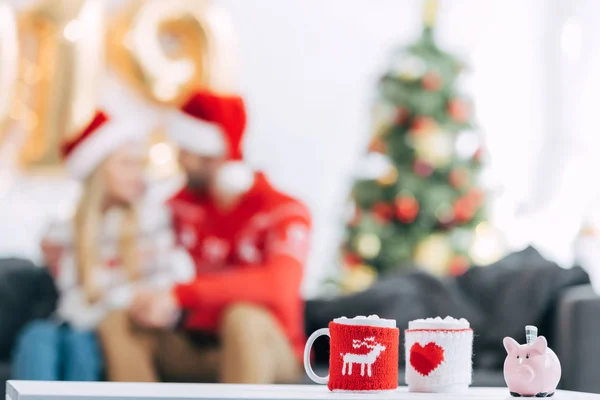 Christmas cocoa cups with marshmallow and piggy bank with banknote on table, couple sitting behind — Stock Photo