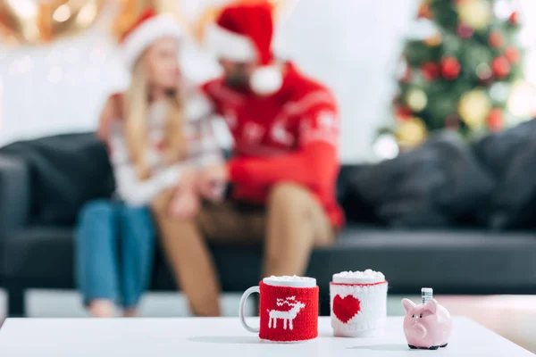 Tazas de café de Navidad y alcancía con billete en la mesa, pareja sentada detrás - foto de stock