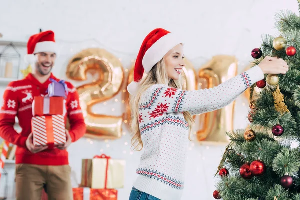 Mujer atractiva decorando el árbol de Navidad mientras que el hombre feliz lleva cajas de regalo - foto de stock