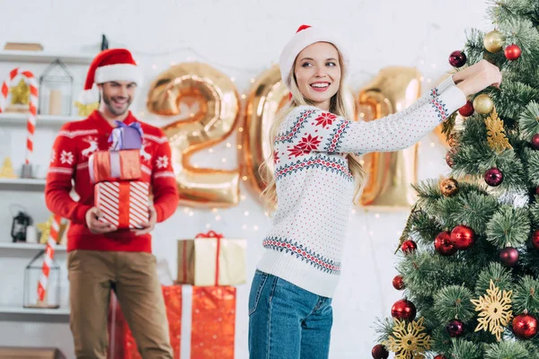 Glückliche Frau schmückt Weihnachtsbaum, während Mann Geschenke trägt — Stockfoto
