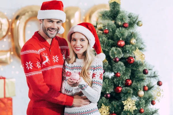 Glückliches Paar in Weihnachtsmützen hält Sparschwein mit Geldschein zu Hause mit Weihnachtsbaum — Stockfoto