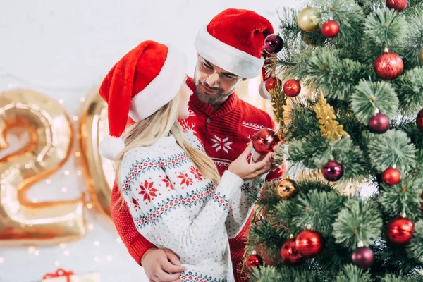 Счастливая пара в шляпах Санты, украшающая рождественскую елку дома вместе — стоковое фото