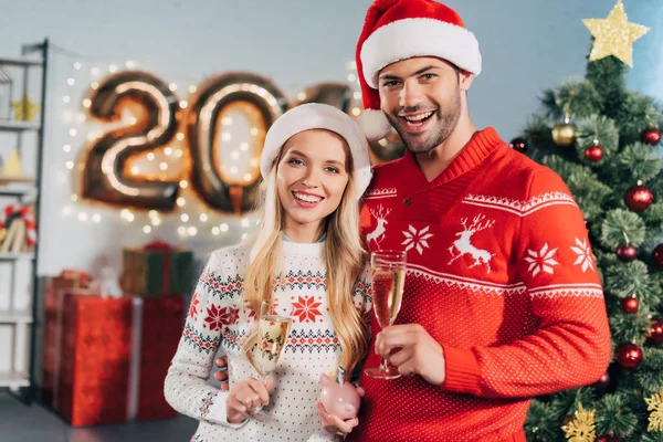 Casal sorrindo com taças de champanhe segurando porquinho banco com poupança para o ano novo — Fotografia de Stock