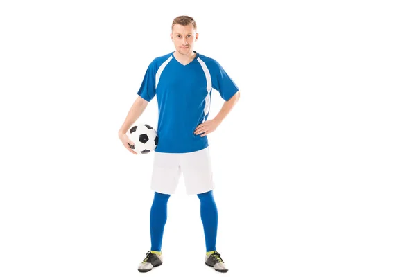 Selbstbewusster junger Fußballer, der Ball hält, während er mit der Hand auf der Taille steht und isoliert auf weiß in die Kamera schaut — Stockfoto