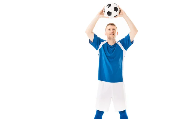 Sonriente joven futbolista sosteniendo la pelota por encima de la cabeza y mirando hacia arriba aislado en blanco - foto de stock