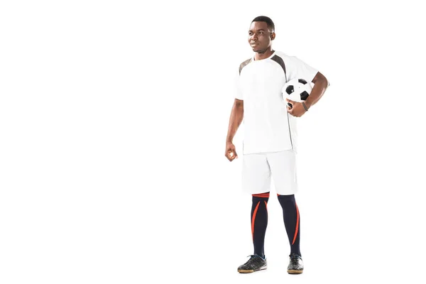 Vista completa de sonriente joven afroamericano futbolista sosteniendo la pelota y mirando hacia otro lado aislado en blanco - foto de stock