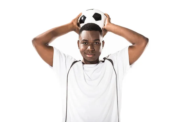 Joven jugador de fútbol afroamericano sosteniendo la pelota por encima de la cabeza y sonriendo a la cámara aislada en blanco - foto de stock