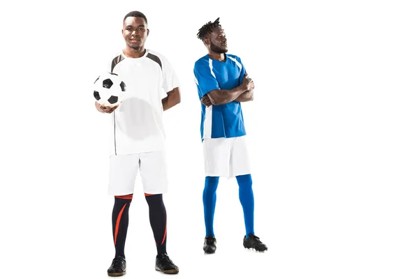 Vista completa del joven jugador de fútbol afroamericano sosteniendo la pelota y sonriendo a la cámara mientras deportista con los brazos cruzados de pie detrás - foto de stock