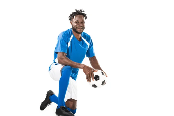 Alegre joven afroamericano jugador de fútbol sosteniendo la pelota y sonriendo a la cámara aislado en blanco - foto de stock