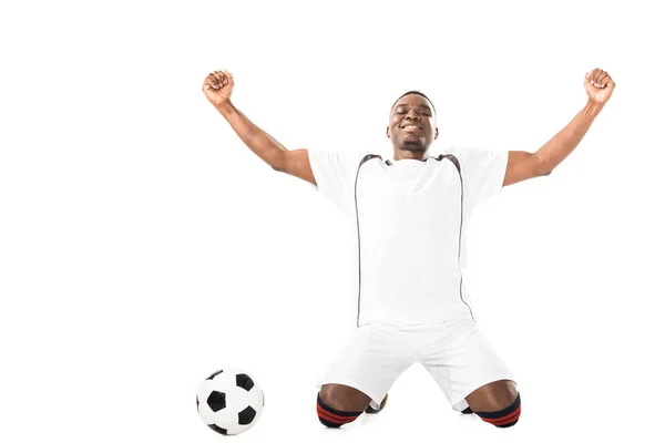 Счастливый молодой триумфальный африканский футболист, стоящий на коленях рядом с мячом, изолированным на белом — стоковое фото