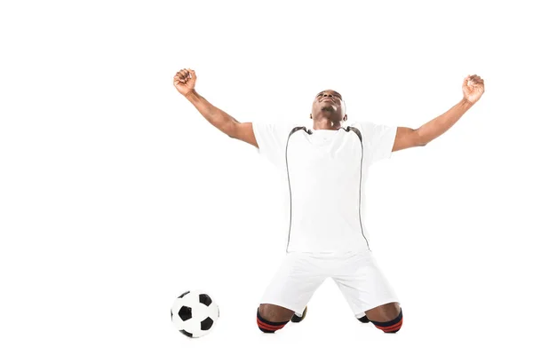 Торжествующий молодой африканский футболист, стоящий на коленях возле изолированного на белом мяче — стоковое фото