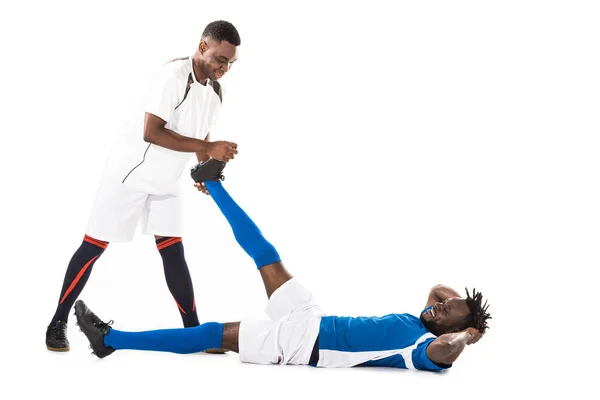 Smiling young african american sportsman stretching leg of soccer player lying isolated on white — Stock Photo