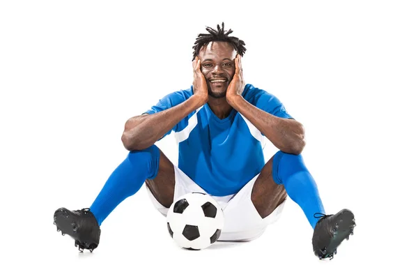 Feliz joven afroamericano jugador de fútbol sentado con la pelota y sonriendo a la cámara aislado en blanco - foto de stock