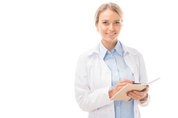 Glückliche junge Ärztin schreibt in Notizbuch und blickt auf Kamera isoliert auf weiß — Stockfoto