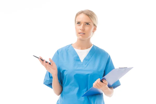 Enfermera joven reflexiva con portapapeles y pluma mirando hacia arriba aislado en blanco - foto de stock
