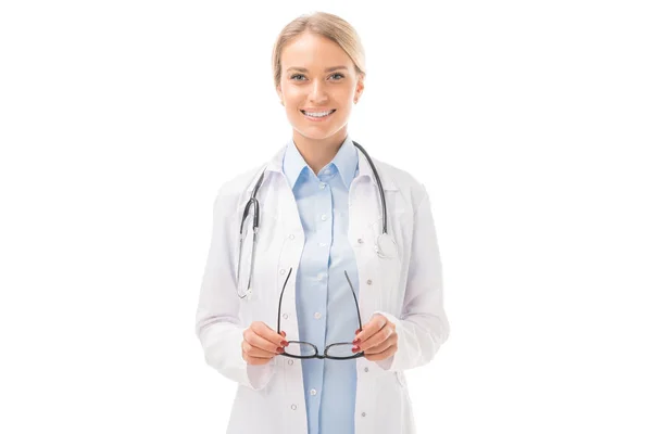 Hermosa joven doctora en abrigo blanco sosteniendo anteojos y mirando a la cámara aislada en blanco - foto de stock