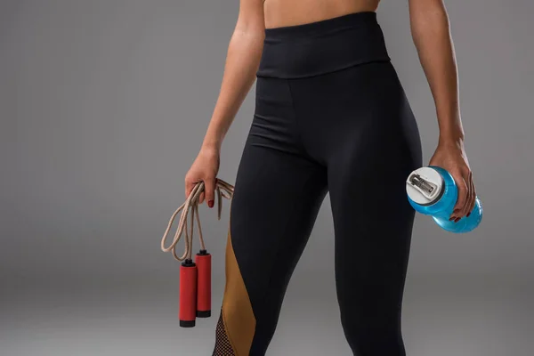 Cropped shot of sportive woman in leggins with jumping rope and water bottle on grey — Stock Photo