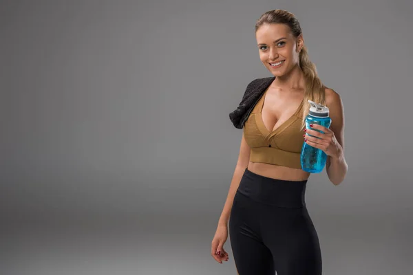 Спортивная молодая женщина с полотенцем и бутылкой фитнес-воды, стоящая на сером и смотрящая в камеру — стоковое фото