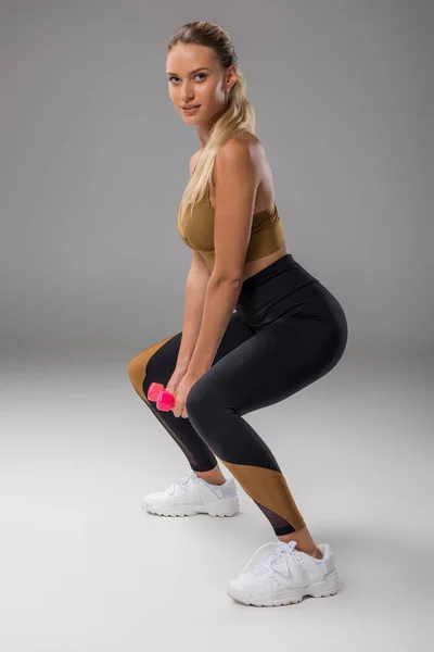 Sportive jeune femme faire squats avec haltères sur gris — Photo de stock