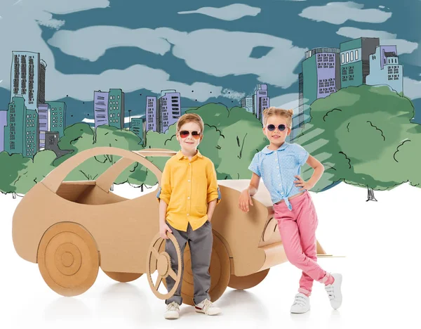 Stylish children in sunglasses posing near cardboard car in drawn city and park — Stock Photo