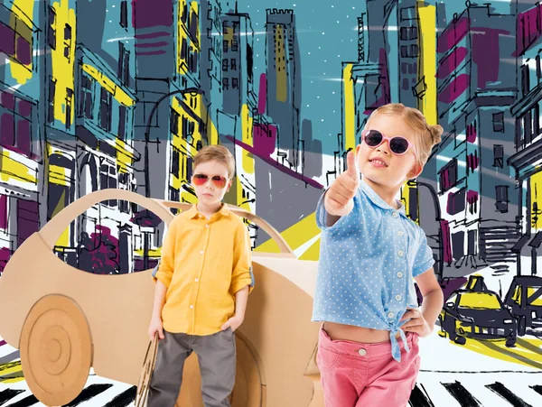 Adorável criança elegante em óculos de sol mostrando polegar para cima enquanto menino de pé perto de carro de papelão na cidade desenhada — Fotografia de Stock