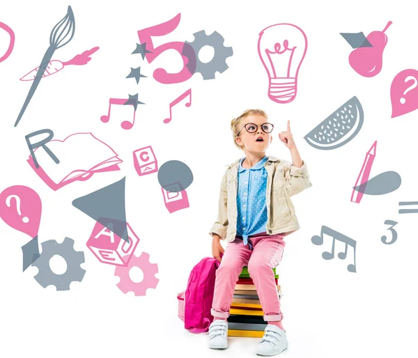Excited schoolchild pointing up and having idea while sitting on pile of books with backpack isolated on white with study icons — Stock Photo